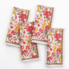 A set of four folded cloth napkins with a floral pattern in red, yellow, and brown colors on a white background, displayed against a neutral background.