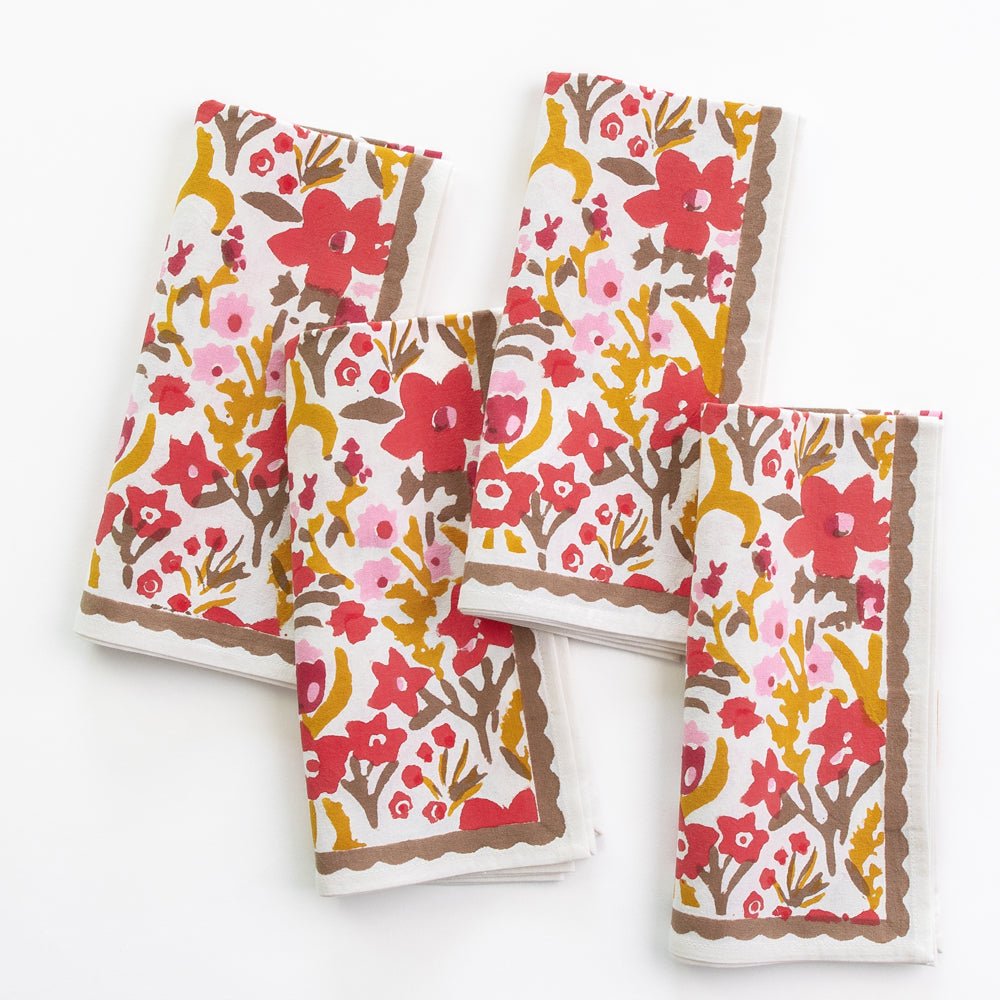A set of four folded cloth napkins with a floral pattern in red, yellow, and brown colors on a white background, displayed against a neutral background.