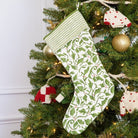 A green and white quilted Christmas stocking with a holly berry print hanging on a Christmas tree.