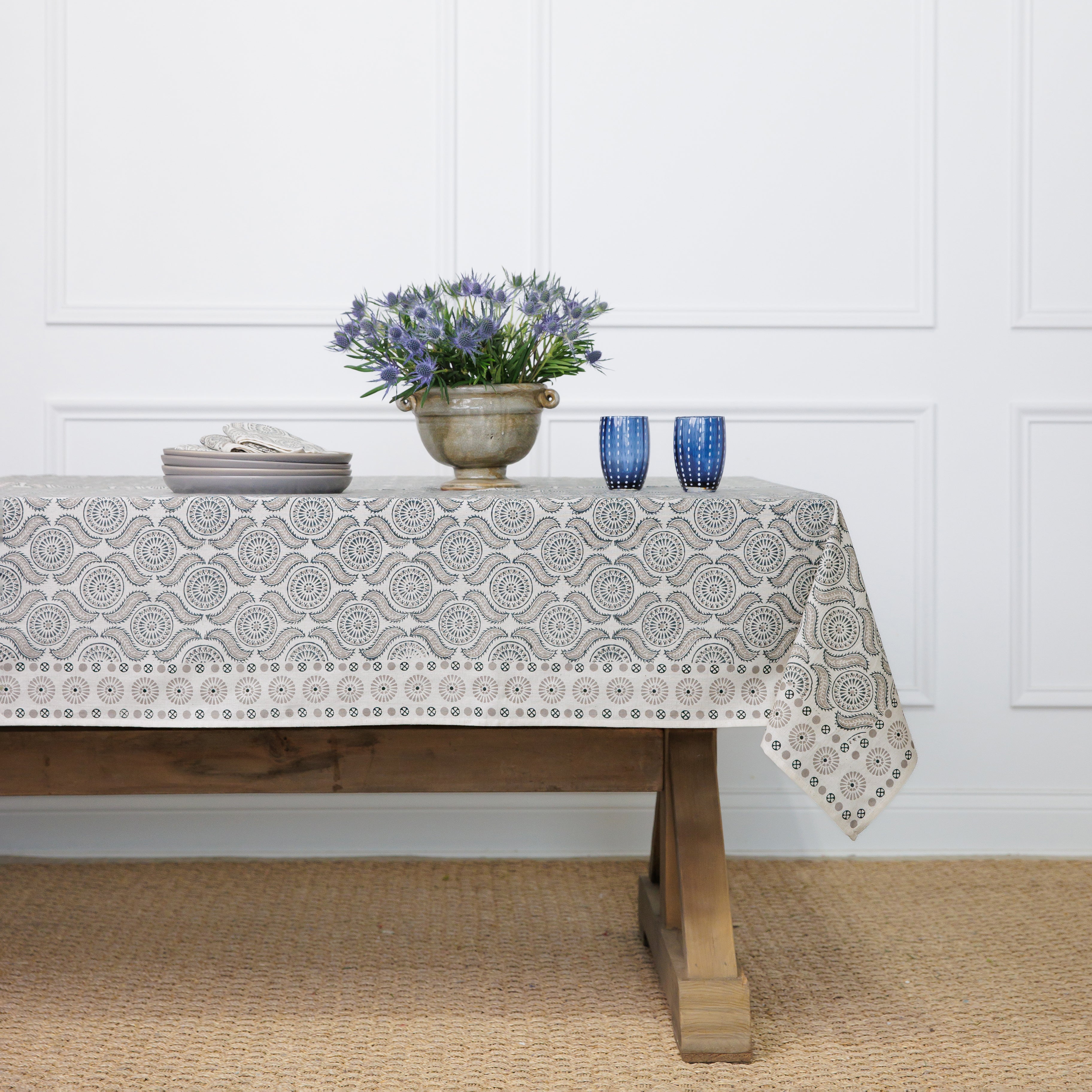 A tablecloth with a geometric pattern featuring medals and mustaches, displayed on a table with a vase of flowers and two blue cups.