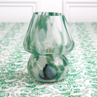 A green and white glass lamp with a conical shade placed on a table with a patterned tablecloth.