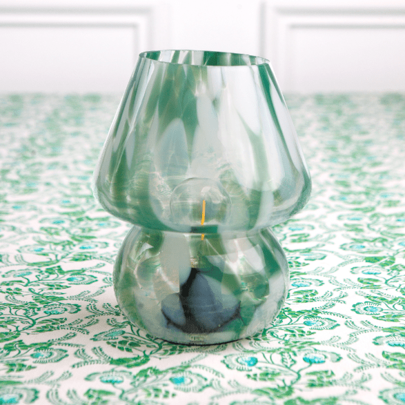 A green and white glass lamp with a conical shade placed on a table with a patterned tablecloth.