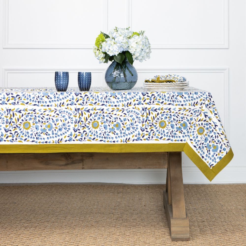 Tablecloth with floral pattern on a wooden table, with a vase of flowers and cups in the background.