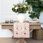 Decorative table runner with floral pattern on a wooden table with a vase of flowers and glasses.