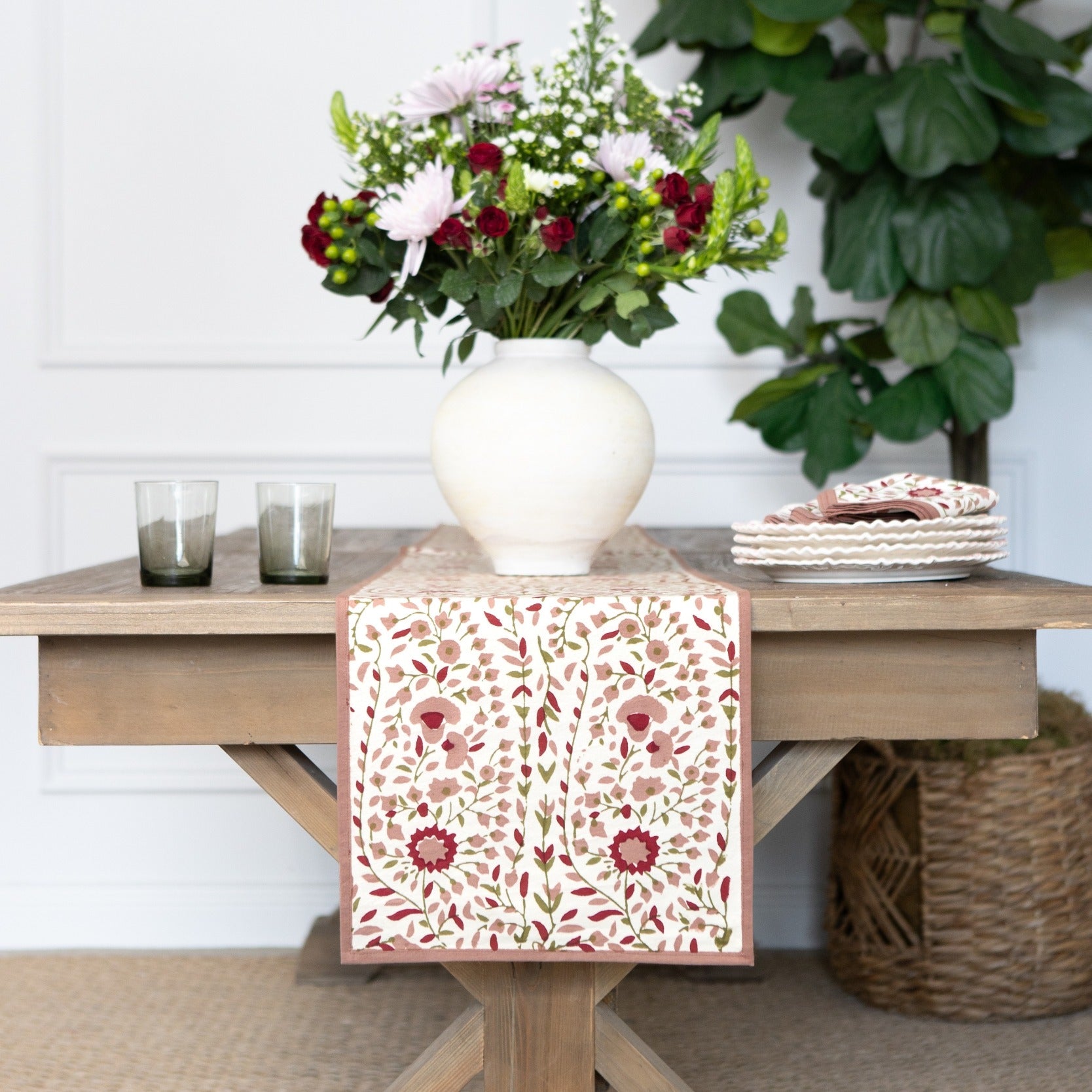 Decorative table runner with floral pattern on a wooden table with a vase of flowers and glasses.