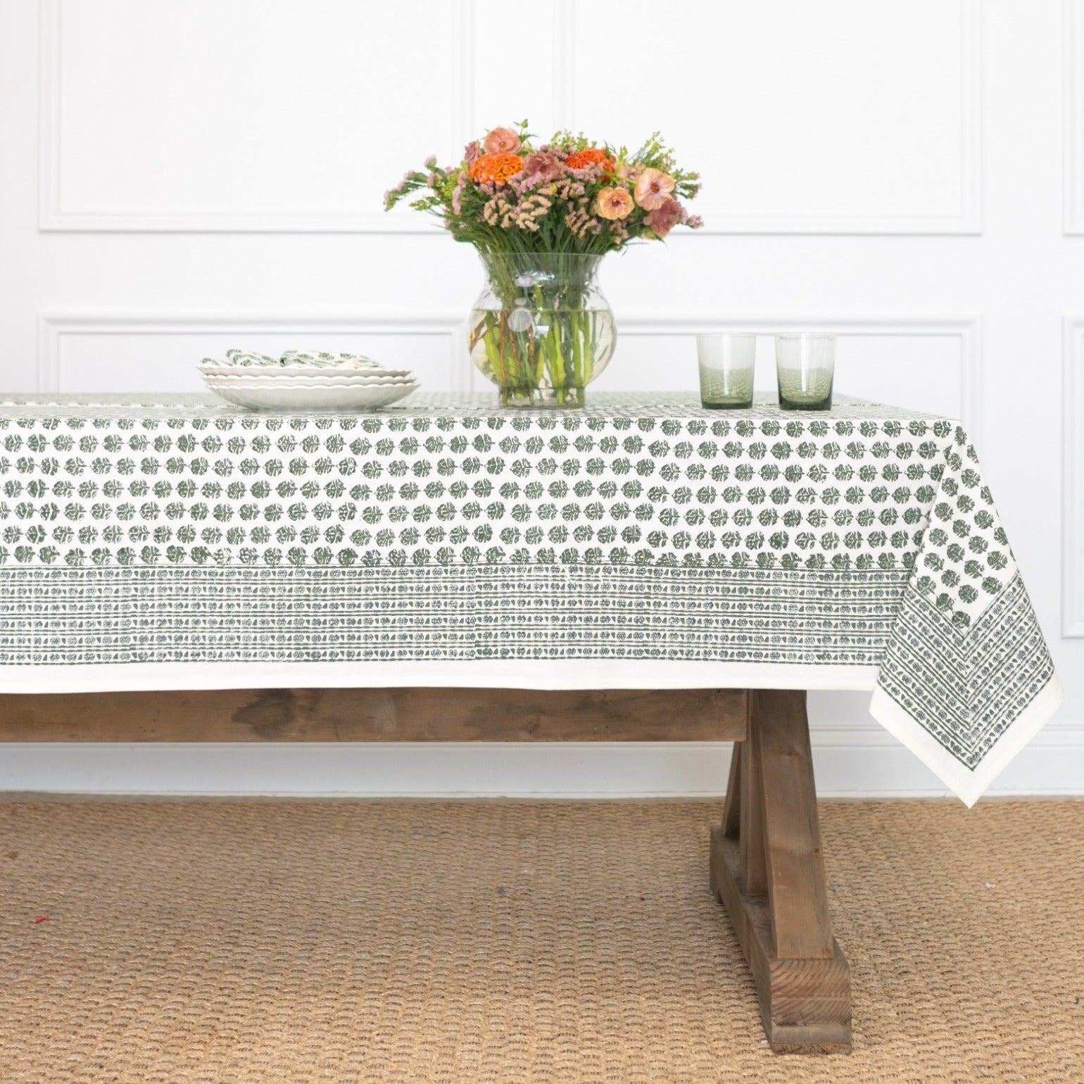 Table with a patterned tablecloth, flowers, and glasses in a room.