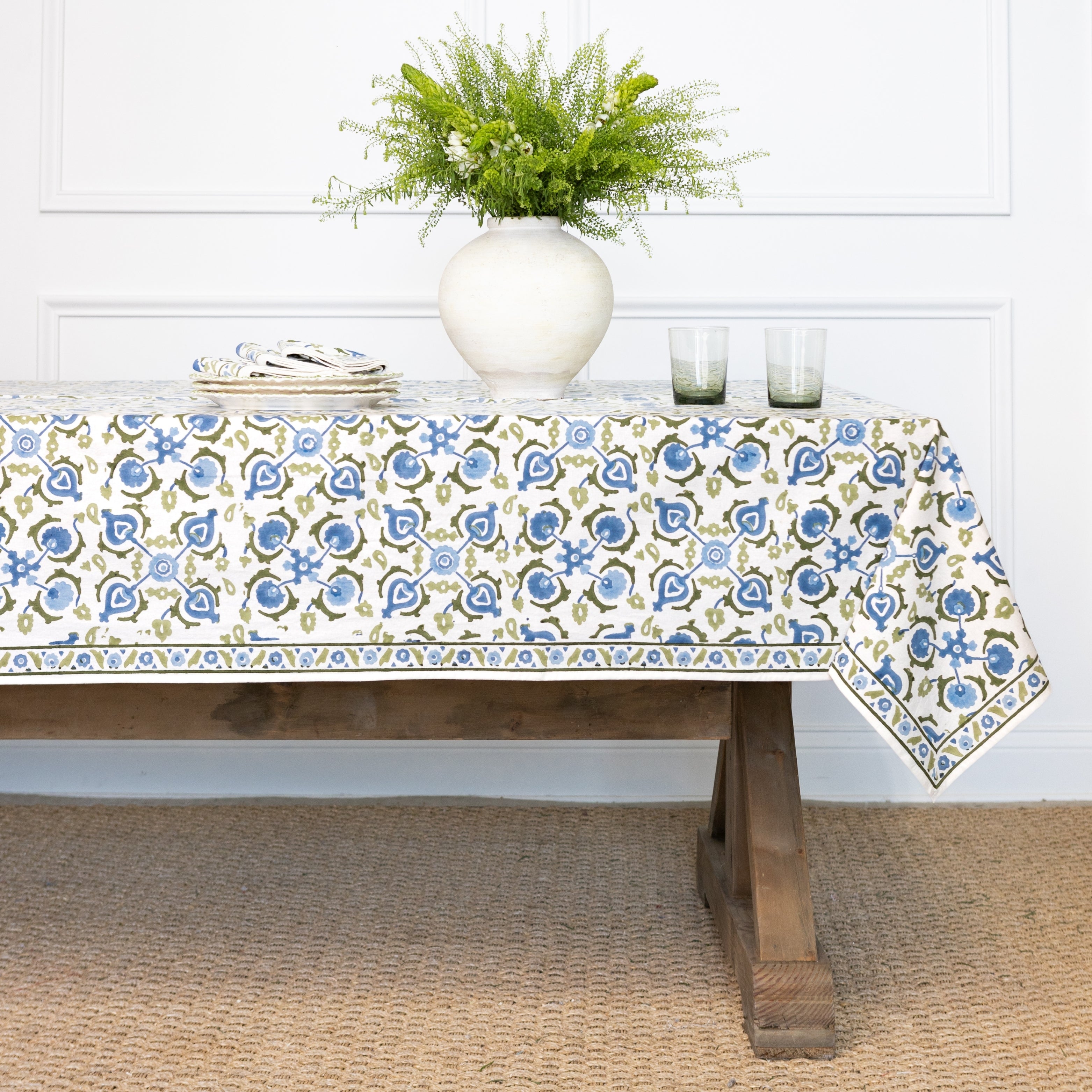 Tablecloth with floral pattern on a wooden table, with a vase of greenery and candles in the background.
