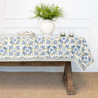 Tablecloth with floral pattern on a wooden table, with a vase of greenery and candles in the background.