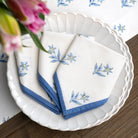 White napkins with blue floral embroidery on a white plate with a floral-patterned tablecloth.