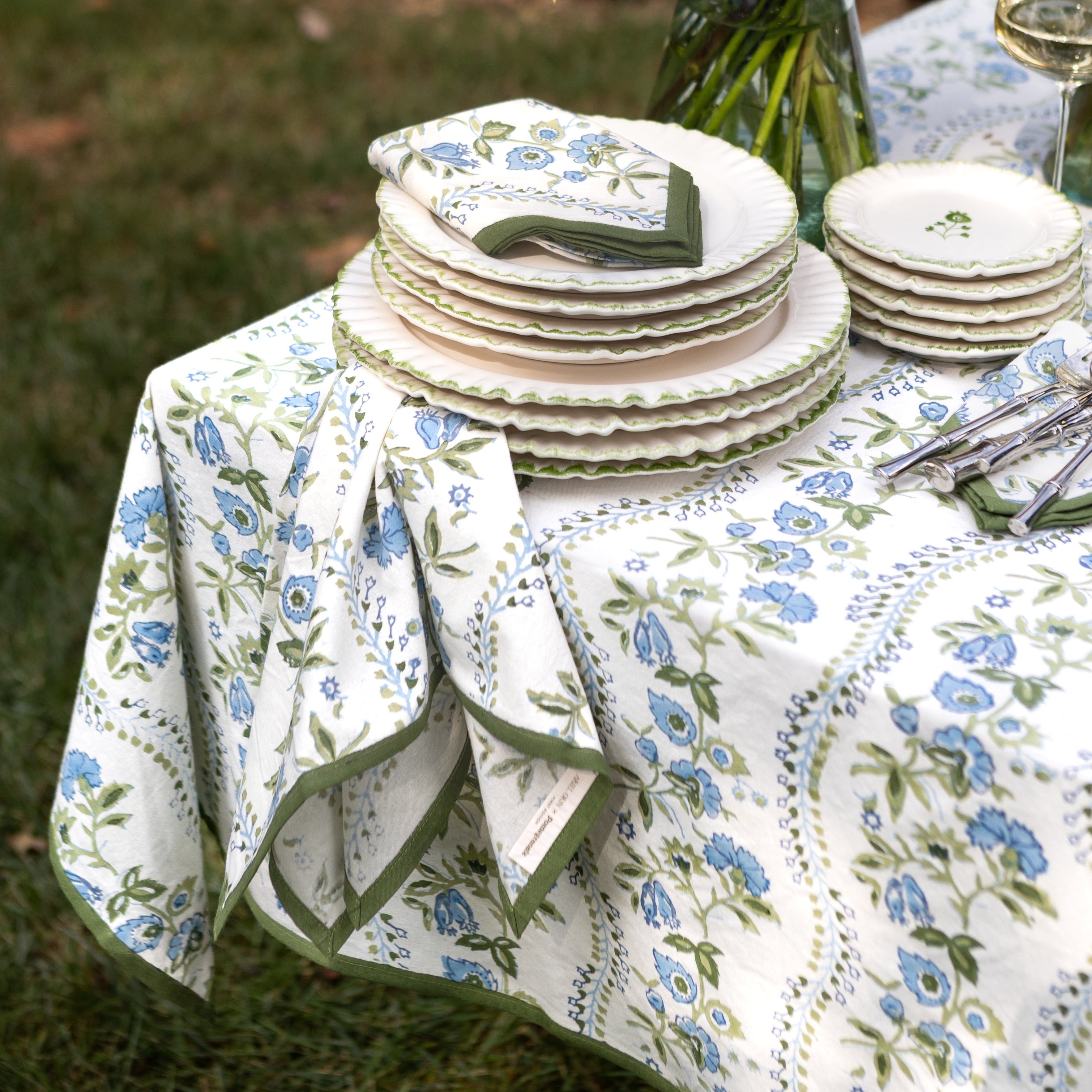Outdoor table setting with floral-patterned tablecloth, plates, and cutlery on a grassy background.