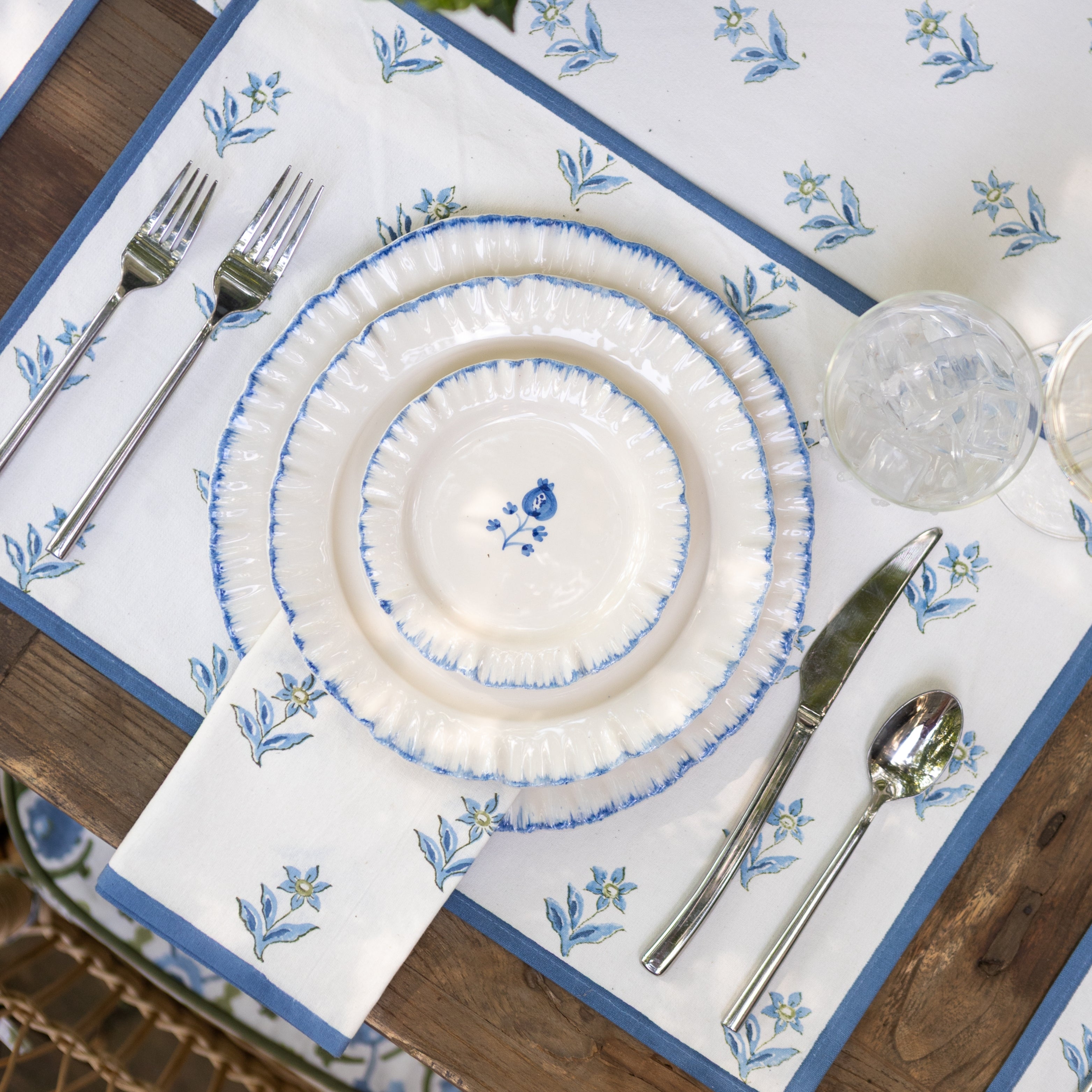 Set table with white plates, silverware, and a decorative blue floral placemat on a wooden table.