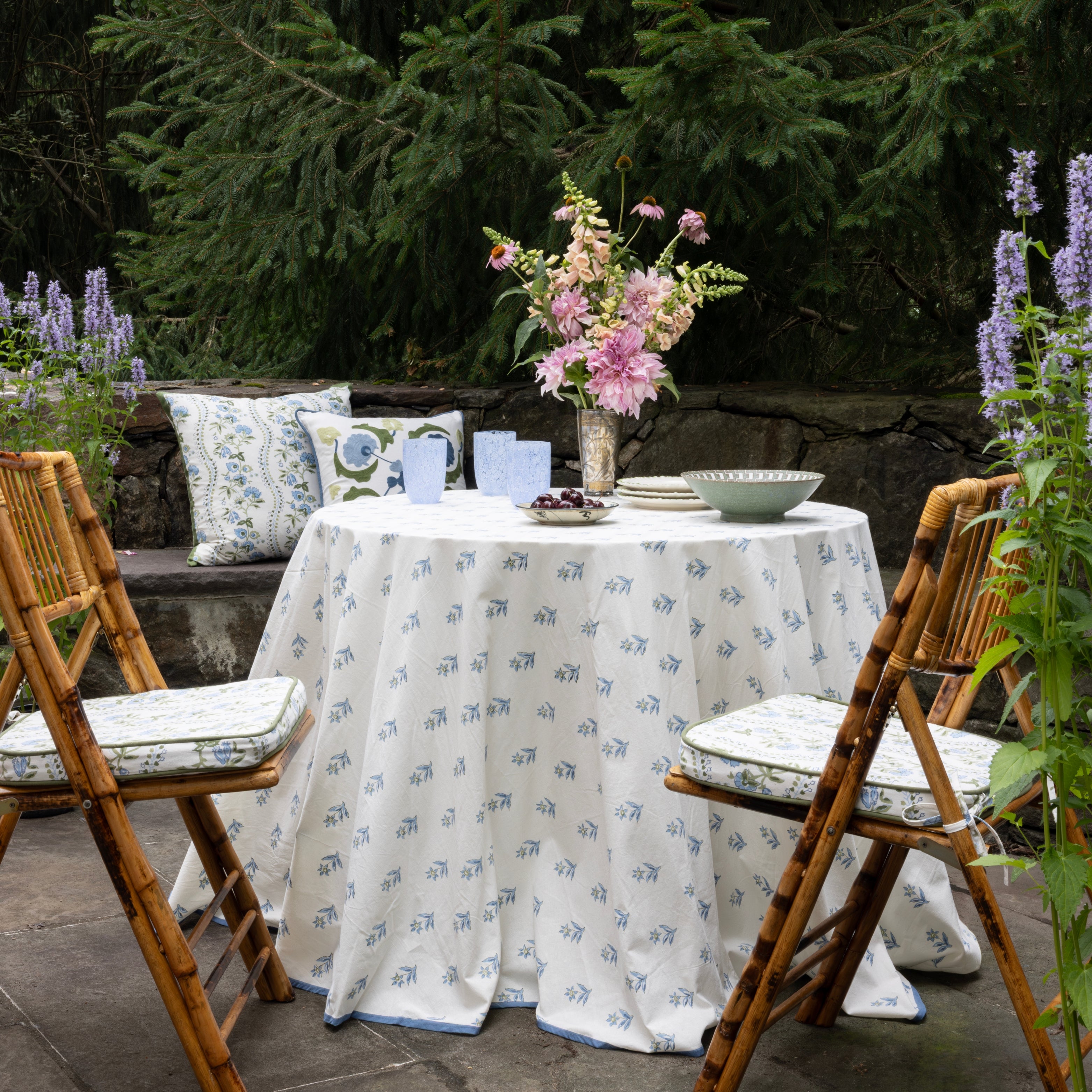 Outdoor setting with a round table covered by a floral-patterned tablecloth, surrounded by wooden chairs with cushions.