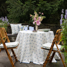 Outdoor setting with a round table covered by a floral-patterned tablecloth, surrounded by wooden chairs with cushions.