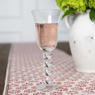 A clear wine glass with a decorative stem, filled with pink liquid, placed on a table with a white and red patterned tablecloth.