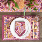 Table setting with pink floral placemat, white plate, gold cutlery, and pink napkin on a wooden table.