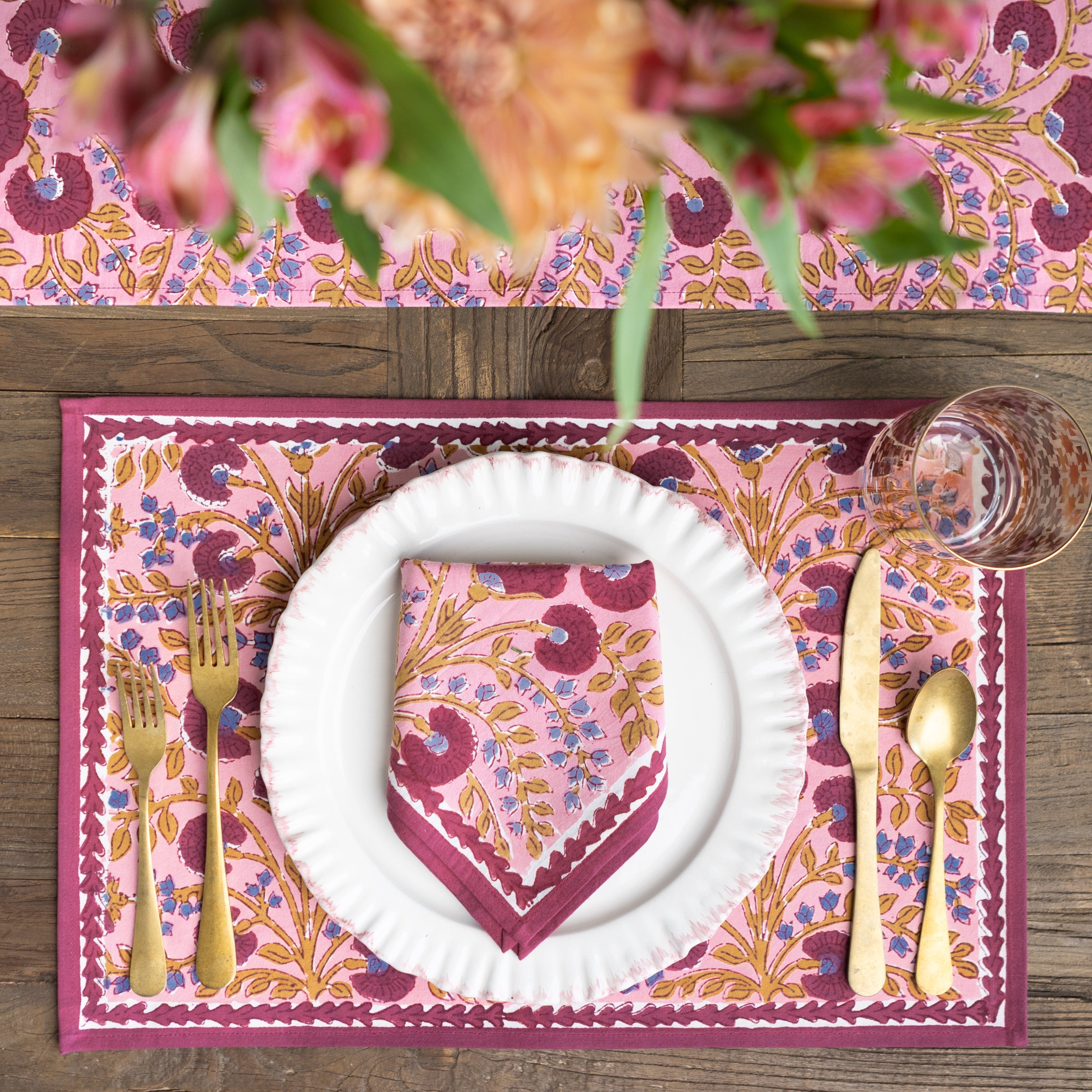 Table setting with pink floral placemat, white plate, gold cutlery, and pink napkin on a wooden table.