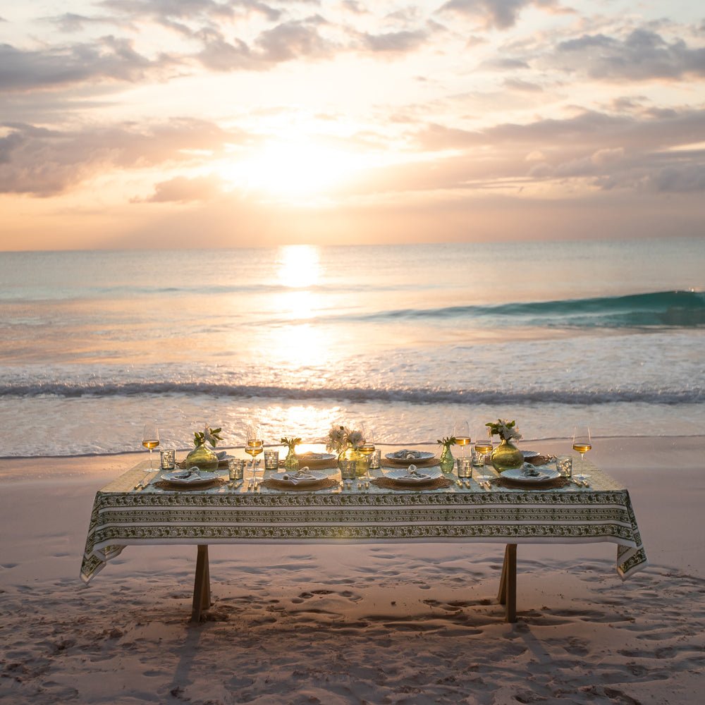 A green and white hand block printed tablecloth on a table placed on a sandy beach with an ocean backdrop.
