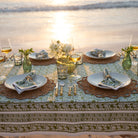 A green and white hand block printed tablecloth on a table placed on a sandy beach with an ocean backdrop.