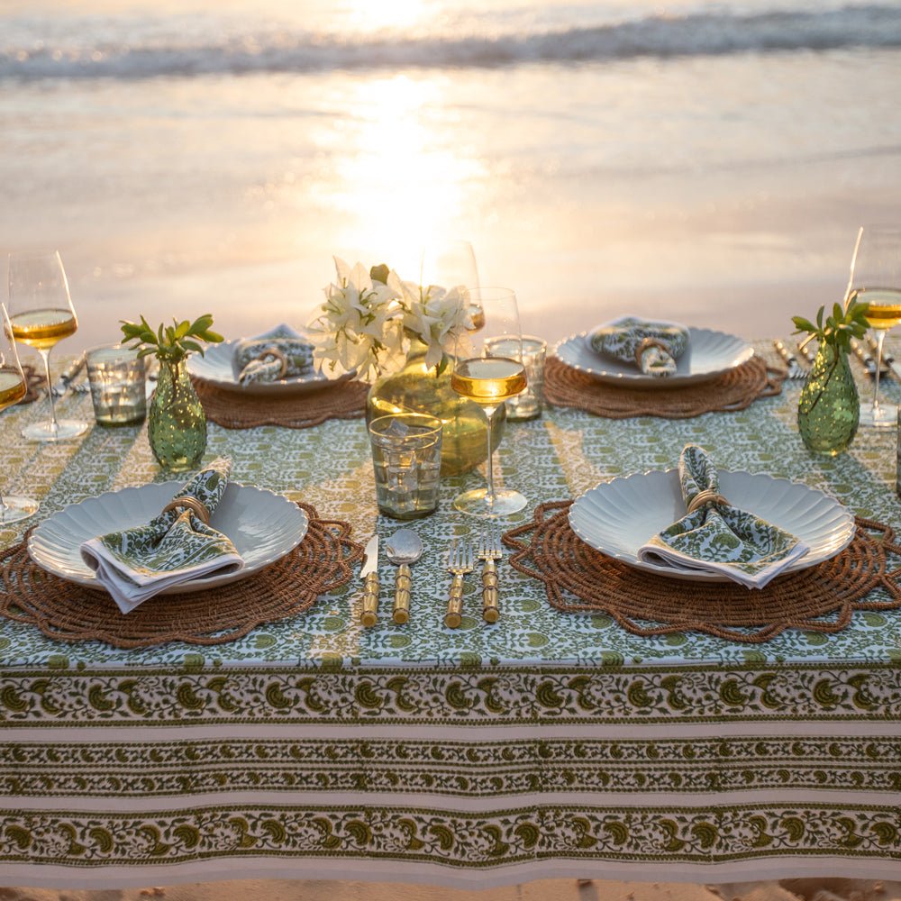 A green and white hand block printed tablecloth on a table placed on a sandy beach with an ocean backdrop.