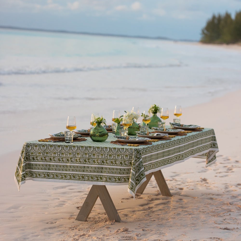 A green and white hand block printed tablecloth on a table placed on a sandy beach with an ocean backdrop.