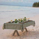 A green and white hand block printed tablecloth on a table placed on a sandy beach with an ocean backdrop.