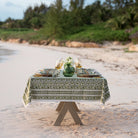 A green and white hand block printed tablecloth on a table placed on a sandy beach with a natural backdrop.
