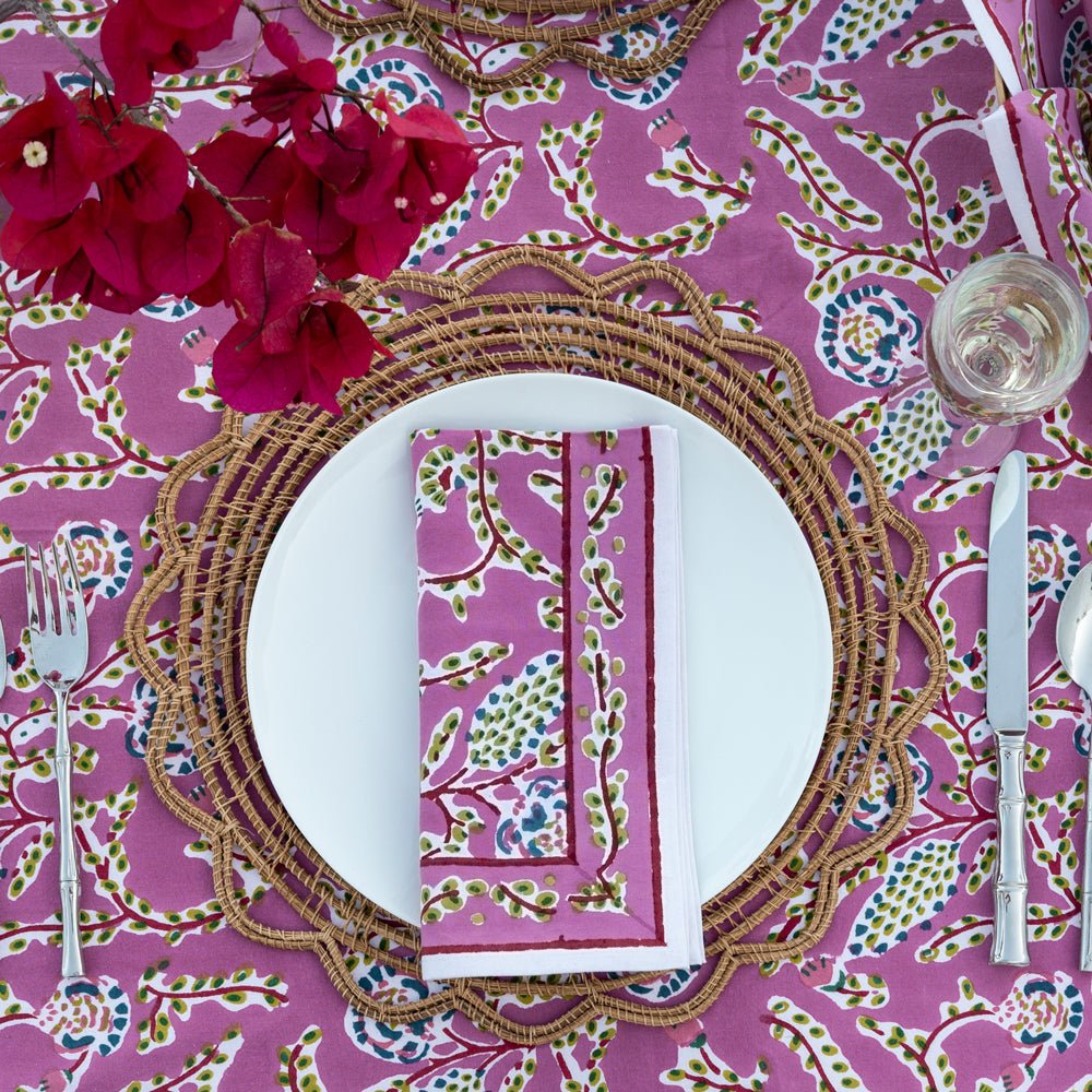 A set of four lilac floral patterned napkins on a dining table with cutlery and a pink floral centerpiece.
