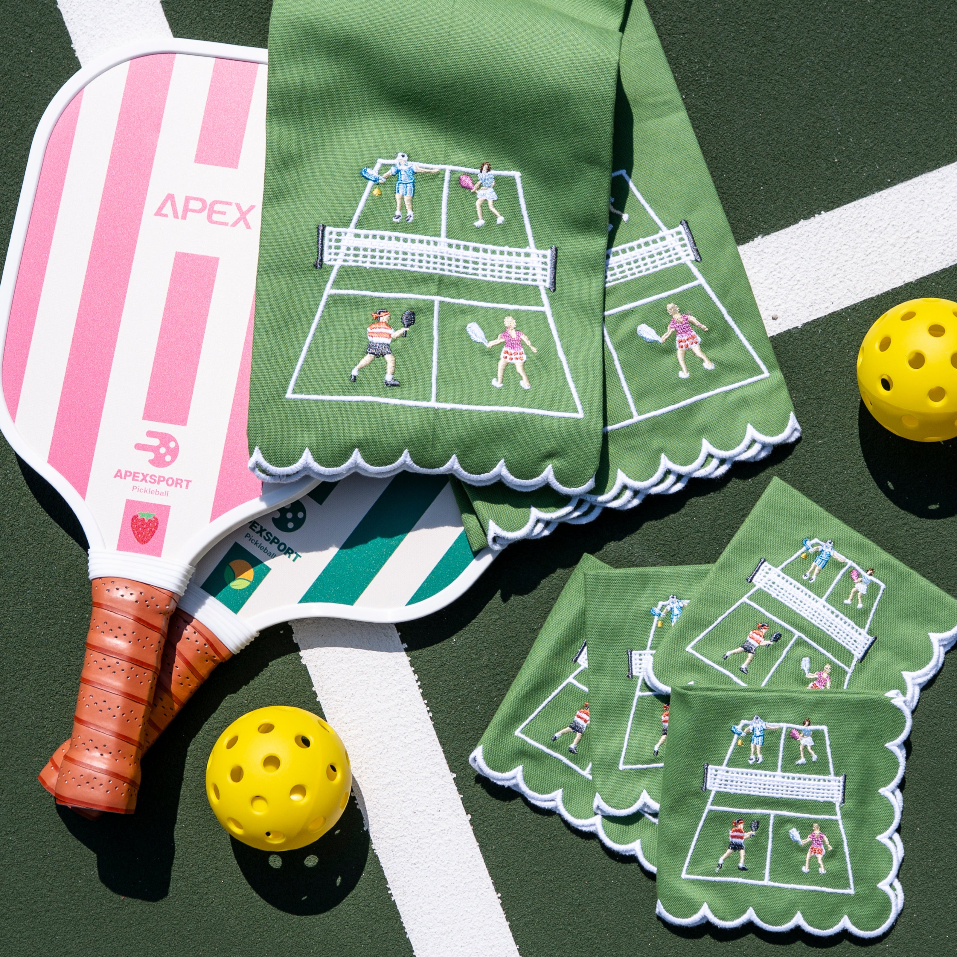 Pickleball paddle with pink and white design, green tea towels with pickleball court illustration, and yellow balls on a green surface.