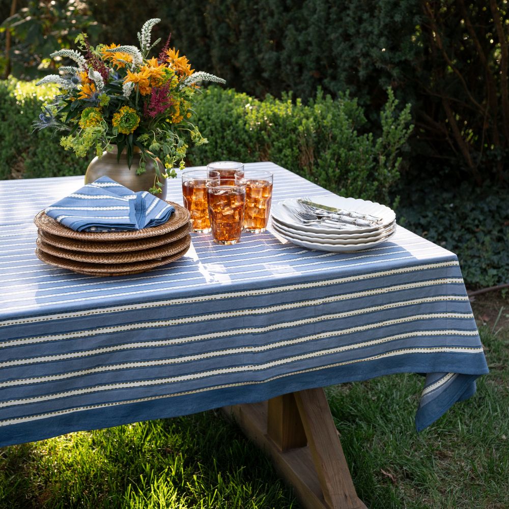 Outdoor table setting with a blue and white striped tablecloth, plates, glasses, and a floral arrangement.
