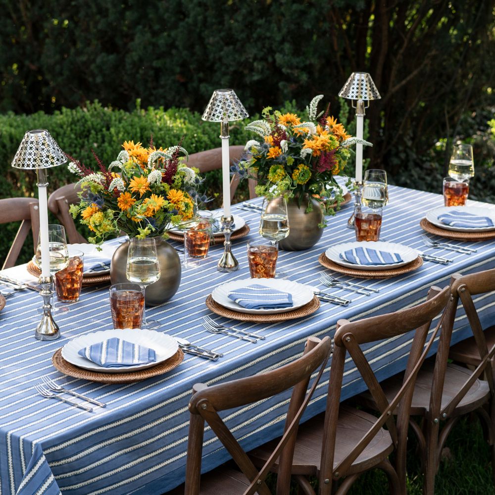 Outdoor dining table set with striped tablecloth, floral centerpieces, and wooden chairs.