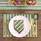 Table setting with a green and white striped placemat, white plate, gold cutlery, and a vase of flowers.