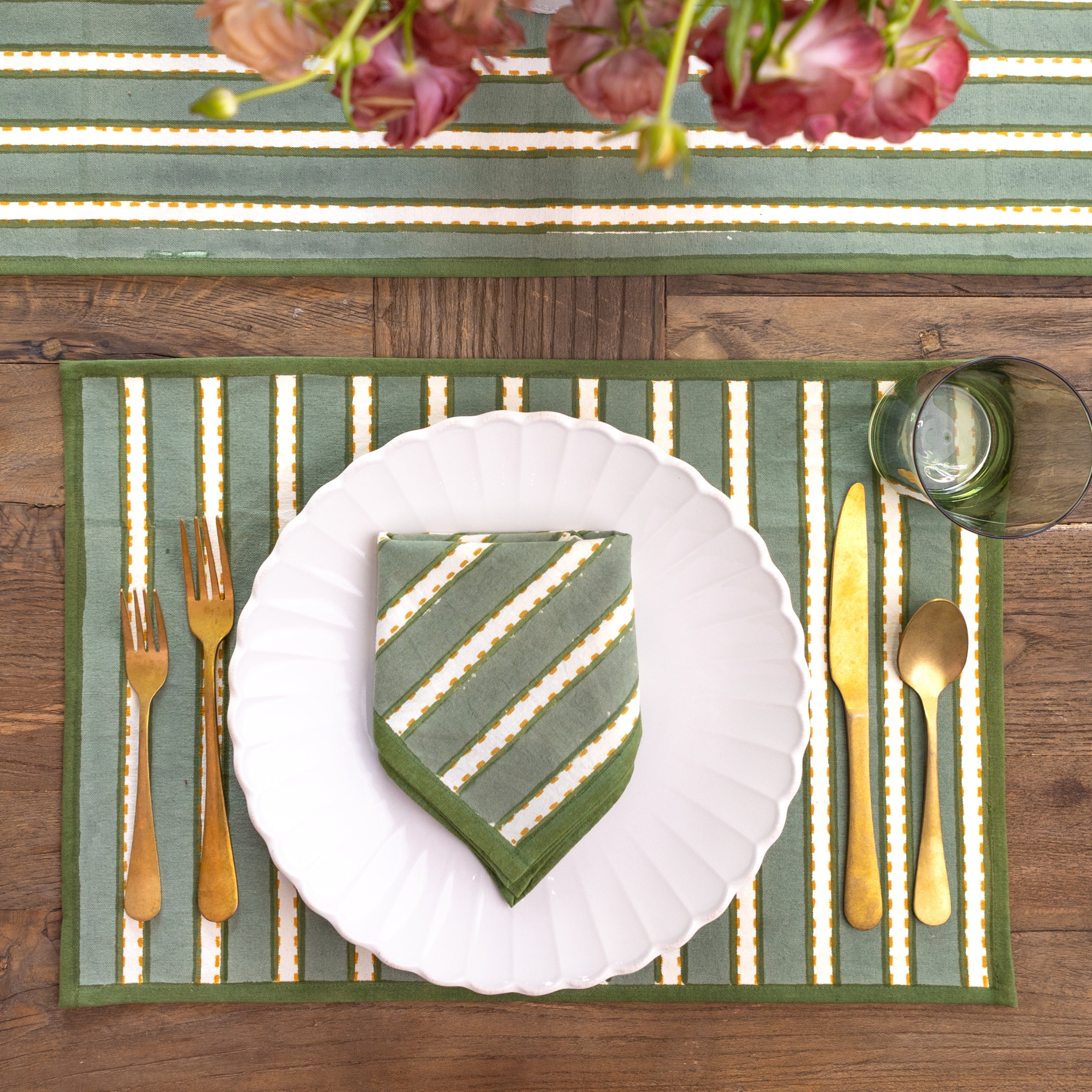 Table setting with a green and white striped placemat, white plate, gold cutlery, and a vase of flowers.