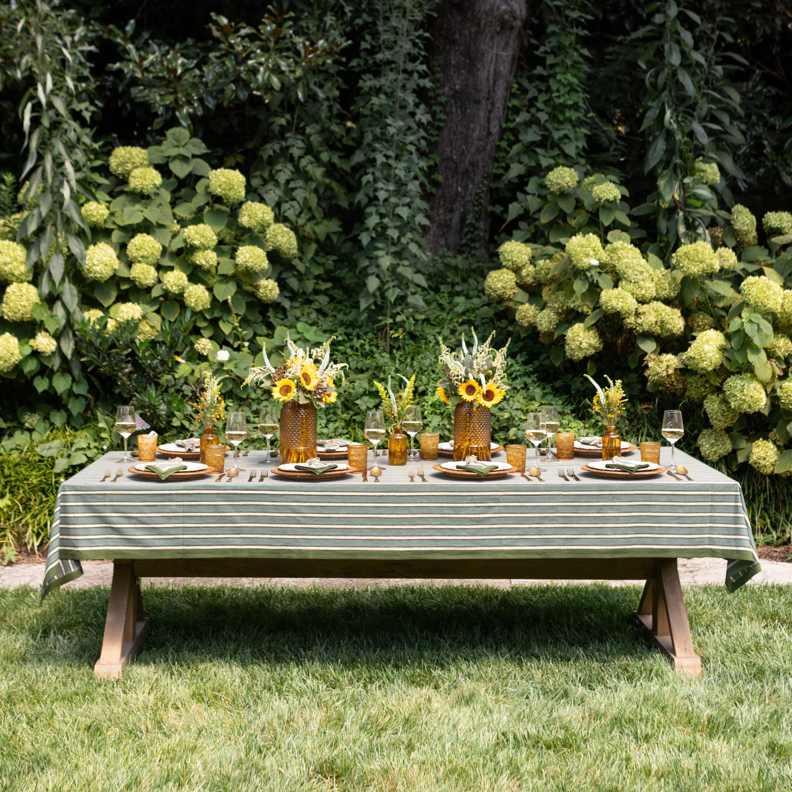 Dining table set for a meal outdoors with a striped tablecloth, sunflowers, and greenery.