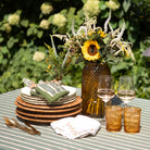 Outdoor table setting with plates, glasses, and a vase of sunflowers on a striped tablecloth.