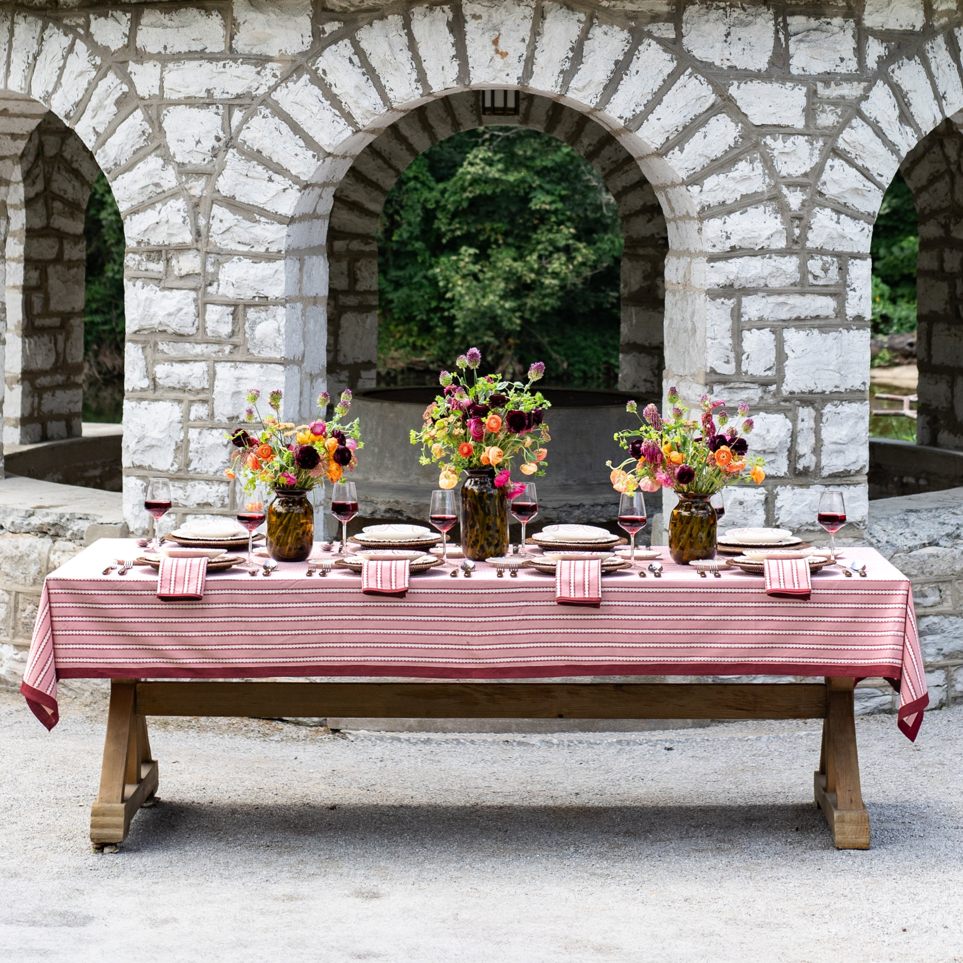 Dining table set with a red tablecloth, floral centerpieces, and wine glasses in front of stone arches.