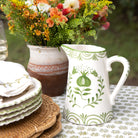 Decorative ceramic pitcher with green patterns on a table with floral arrangement and plates.