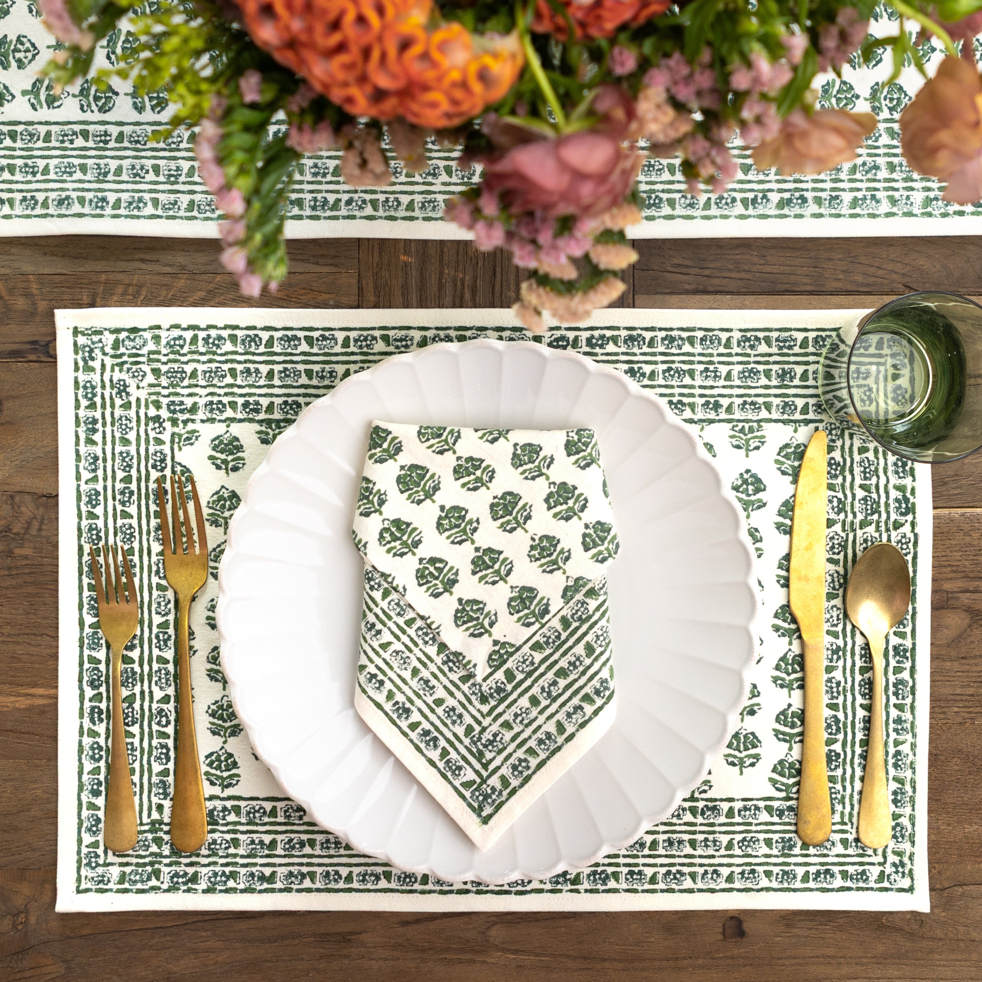 Table setting with a green patterned placemat, white plate, and gold cutlery on a wooden table.
