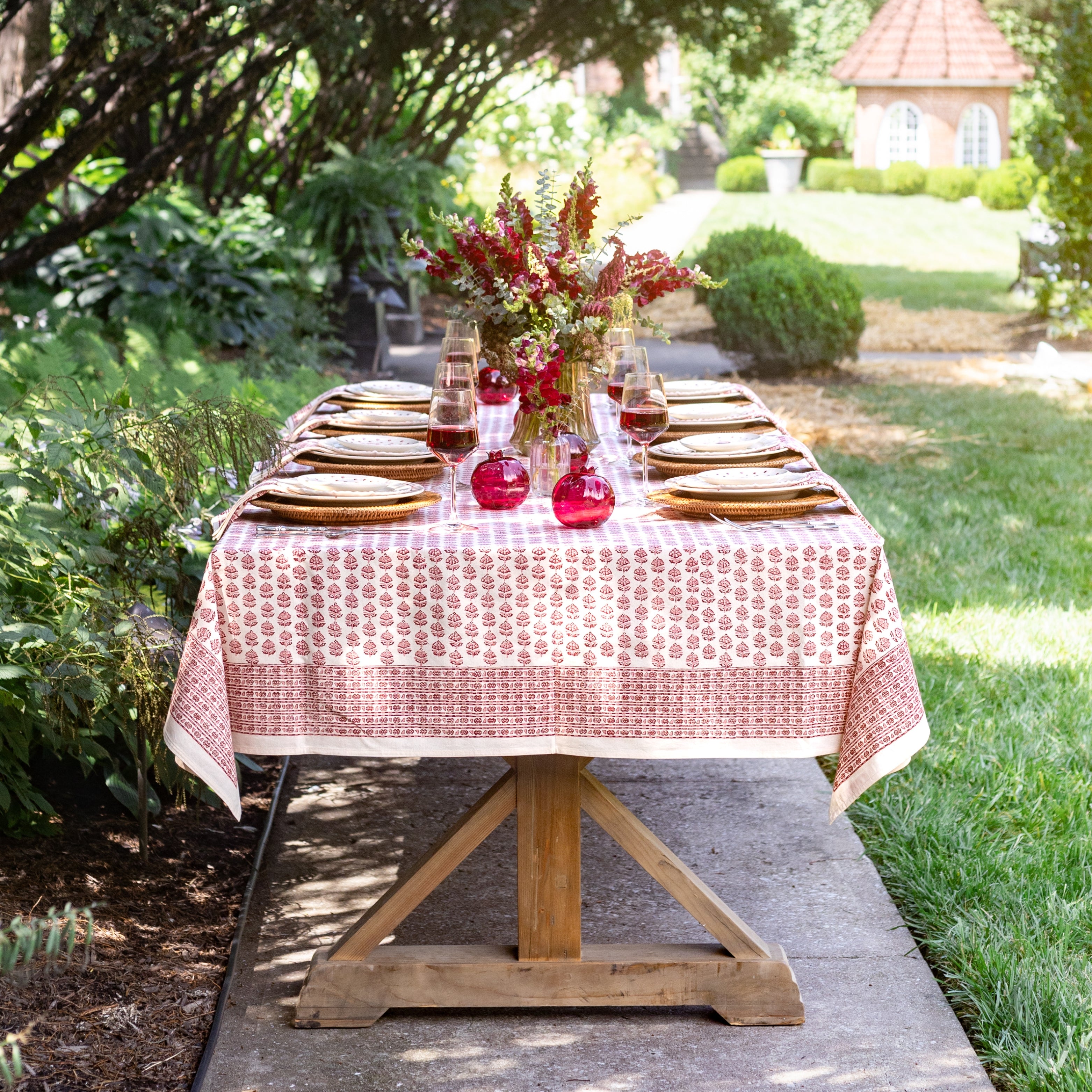 Dining table set outdoors with a floral arrangement and red vases on a red patterned tablecloth.