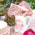 White pitcher with red floral pattern on a table with matching red and white patterned napkin.