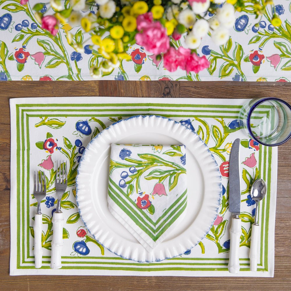 A set of four placemats with a floral pattern in shades of yellow, blue, and red, displayed on a table with a plate and cutlery.