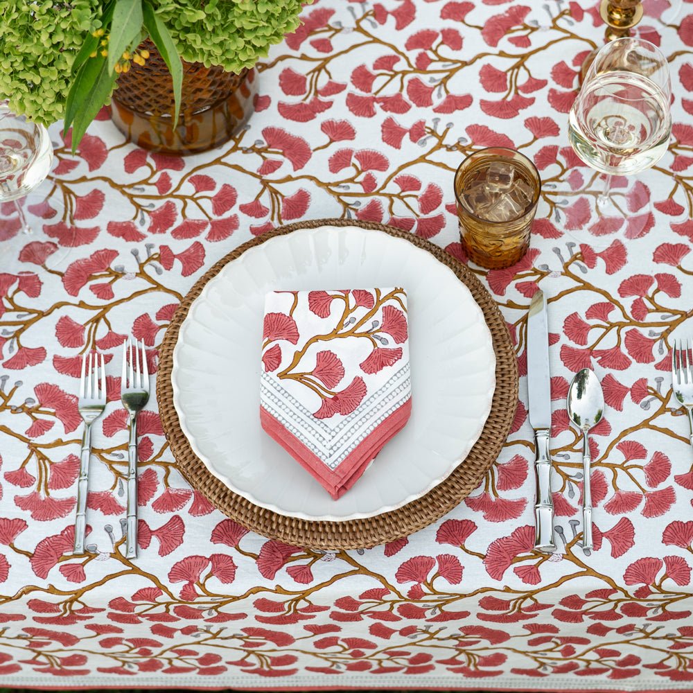 A table setting featuring a folded cotton napkin with a ginkgo leaf pattern in coral hues, displayed on a matching tablecloth