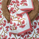 A set of folded cotton napkins with a ginkgo leaf pattern in coral hues, displayed on a woven charger.