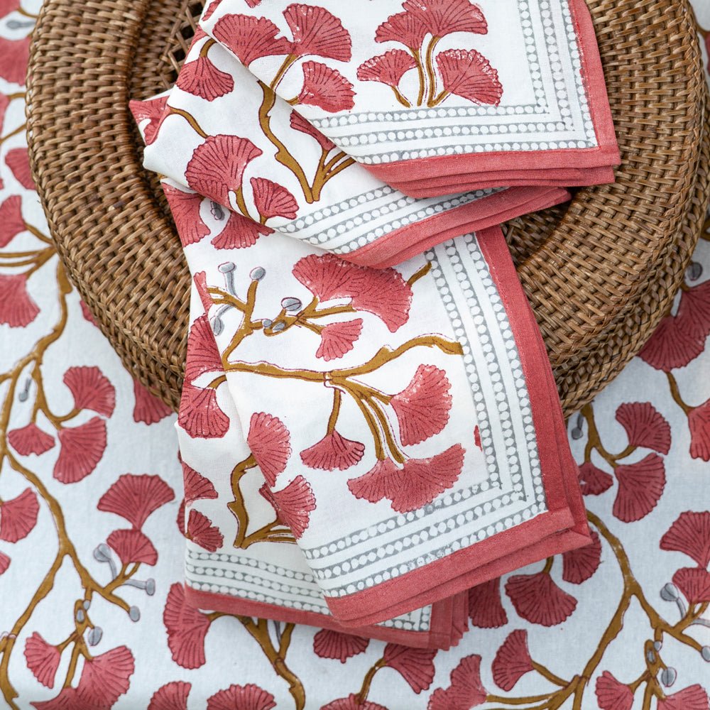 A set of folded cotton napkins with a ginkgo leaf pattern in coral hues, displayed on a woven charger.