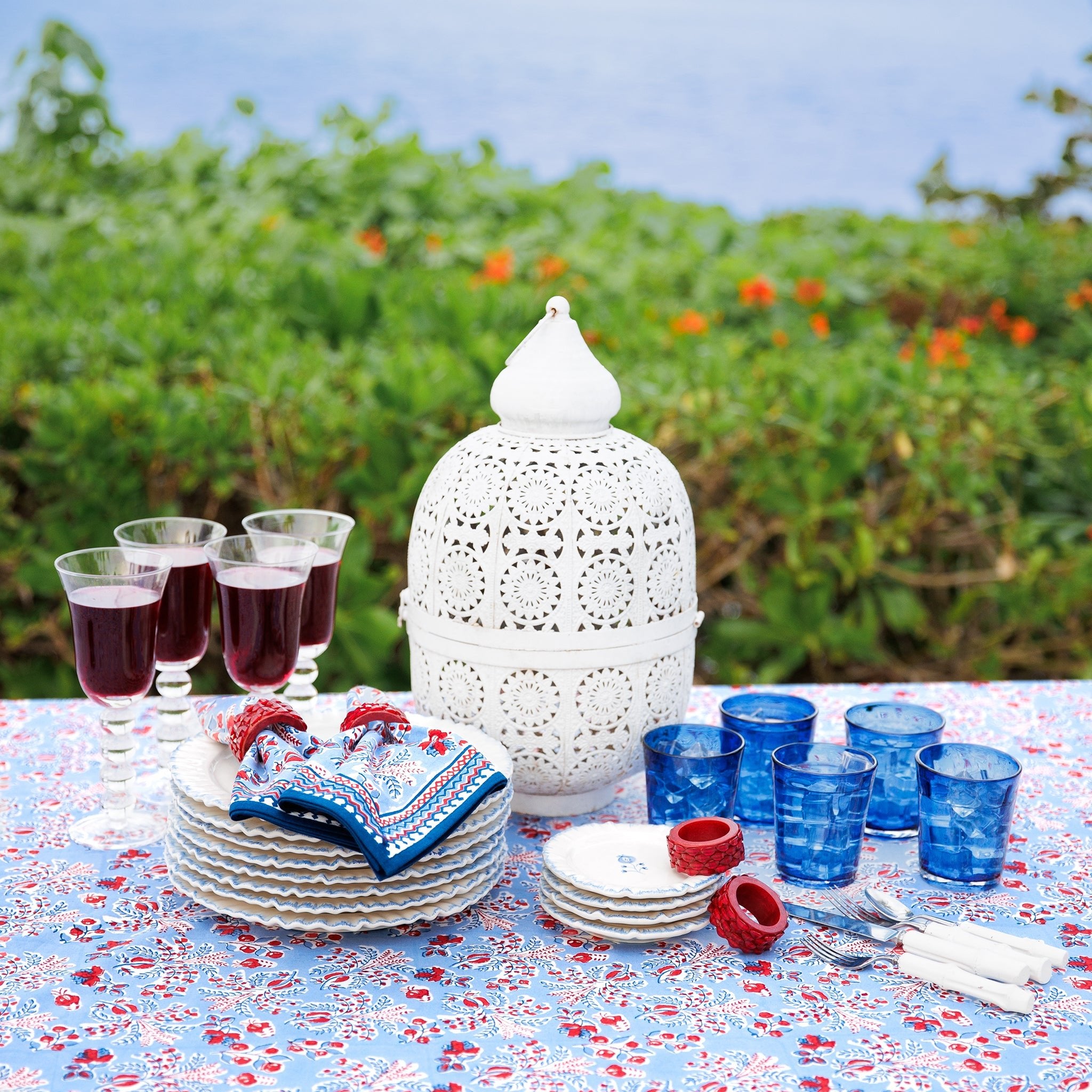 Outdoor table setting with napkins, white lantern, glasses, and plates on a floral tablecloth.