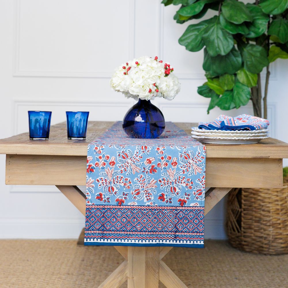 Table with a floral table runner, blue glasses, and a vase with white flowers.