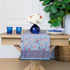 Table with a floral table runner, blue glasses, and a vase with white flowers.