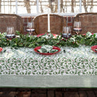 A set table covered with a green and white tablecloth featuring a holly berry pattern, arranged with dishes and silverware.