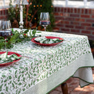 A set table covered with a green and white tablecloth featuring a holly berry pattern, arranged with dishes and silverware.