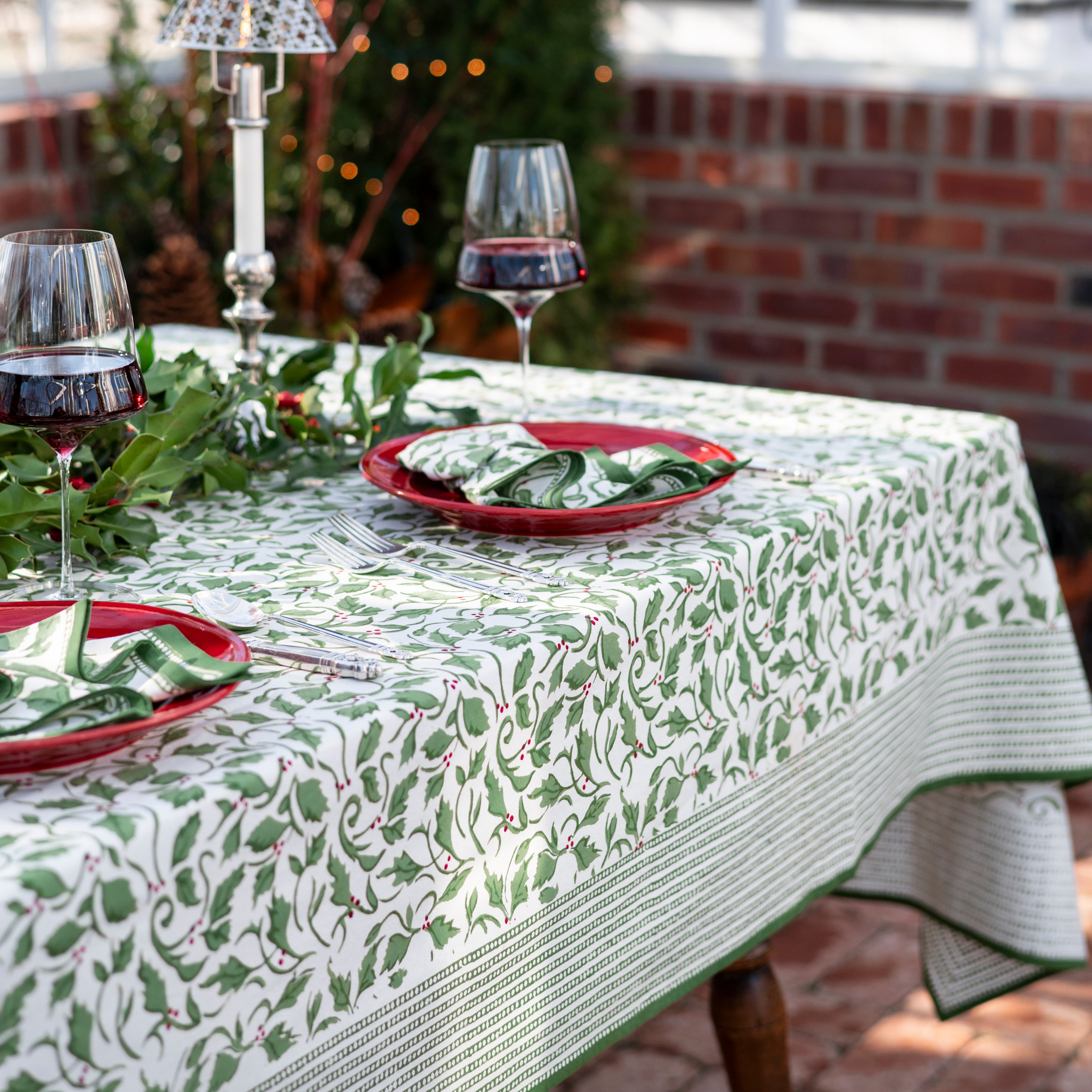 A set table covered with a green and white tablecloth featuring a holly berry pattern, arranged with dishes and silverware.