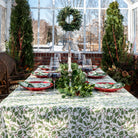 A set table covered with a green and white tablecloth featuring a holly berry pattern, arranged with dishes and silverware.